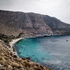 Bahía de aguas turquesas en la playa Cabo de Gata, con barcos navegando y montañas áridas al fondo.