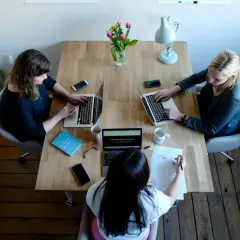 Trzy kobiety pracują przy drewnianym stole, każda z laptopem. Jedna z nich pisze notatki, pokazując, kim jest autorytet w tej grupie.