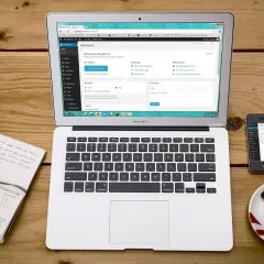 Laptop, notatnik z długopisem, filiżanka kawy i telefon z danymi telefonu na drewnianym blacie.