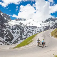 Two cyclists enjoy an epic adventure on a winding road, part of amazing austria bike tours, with snow-capped mountains in the background.