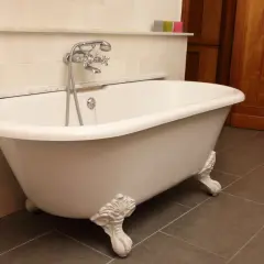 Baignoire îlot blanche sur pieds sculptés, évoquant le poids d'une baignoire en fonte. Robinet argenté, carrelage mural clair et sol sombre.