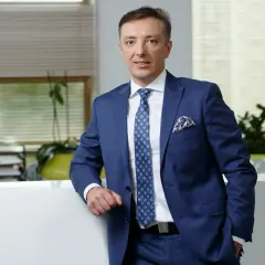 Mężczyzna w granatowym garniturze, białej koszuli i niebieskim krawacie stoi oparty o biały blat.
