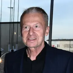 Bogusław Linda w granatowej marynarce i czarnej koszuli, uśmiecha się.