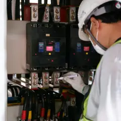 Elektryk w białym kasku i masce zabezpieczającej oznacza śruby przy głównym bezpieczniku prądu.