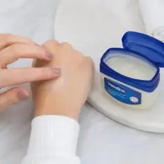 Dłonie rozsmarowują wazelinę techniczną na skórze, która staje się nawilżona i gładka. Słoiczek z produktem stoi obok.
