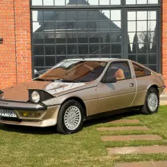 Złoty Talbot Murena z lat 80. na tle ceglanego budynku i zielonej trawy.