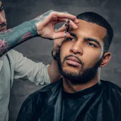 Barbero con tatuajes en el brazo recorta las cejas a un hombre con un corte buzz cut y barba.
