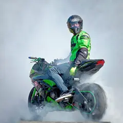 Motociclista con casco y traje verde realiza un caballito, generando humo. La moto, con detalles verdes, parece lista para una demostración de homologación e24.