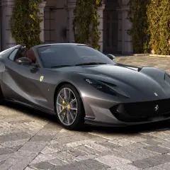 Szary kabriolet Ferrari 812 GTS z żółtymi zaciskami hamulcowymi stoi na brukowanym dziedzińcu przed budynkiem porośniętym bluszczem.