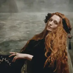 Florence Welch, jak królowa, z rudymi włosami i w czarnej sukni, na tle morskiego krajobrazu.