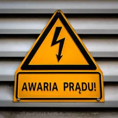 Żółty trójkąt z symbolem pioruna i tabliczka "AWARIA PRĄDU!" na szarej, metalowej powierzchni.