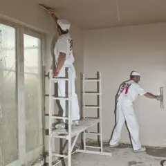 Pracownicy w białych kombinezonach nakładają gładź na tynk cementowo-wapienny. Jeden z nich stoi na podwyższeniu, drugi na drabinie.
