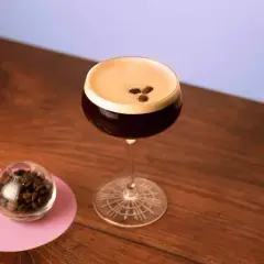 Espresso Martini with coffee beans on top, next to a small glass of coffee beans on a pink coaster.