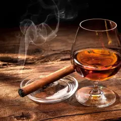 Ein Glas bernsteinfarbener Bourbon mit aufsteigendem Rauch von einer Zigarre auf einem Holztisch.