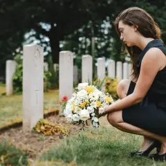Smutna kobieta w czarnej sukience klęczy przy grobie z bukietem kwiatów. W jej oczach widać ból, jakby szukała pocieszenia w cytatach dla zmarłego dziecka.