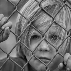 Czarno-białe zdjęcie dziecka z blond włosami, patrzącego przez siatkę ogrodzeniową.