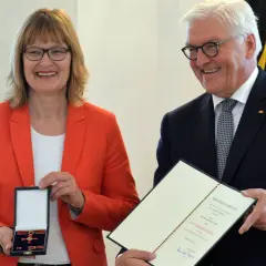 Frau mit rotem Blazer erhält das Bundesverdienstkreuz. Präsident Steinmeier überreicht die Auszeichnung und Urkunde.