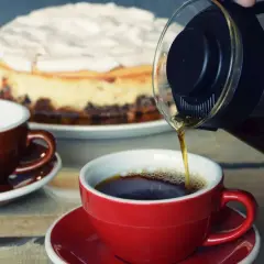 Gorąca woda do kawy nalewa się do czerwonej filiżanki. Obok sernik i brązowa filiżanka.