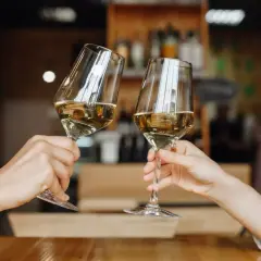 Zwei Hände halten Gläser mit einem goldenen Cuvée Wein. Ein Toast auf den Genuss in gemütlicher Atmosphäre.