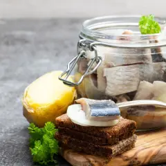Słoik pełen śledzi z cebulką w oleju, obok kromka chleba z kawałkiem śledzia i cebuli, ziemniak i natka pietruszki.