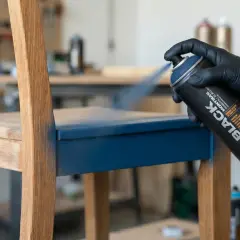 Main d'une personne portant un gant noir pulvérise de la peinture bleue sur un siège de chaise en bois, transformant un meuble mélaminé.