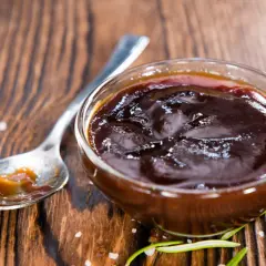 Un cuenco de cristal lleno de deliciosa salsa barbacoa casera, con una cuchara al lado y romero fresco.