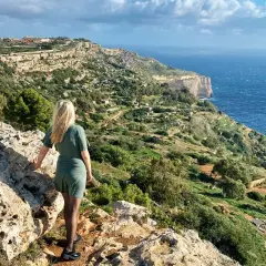 Malta, luty. Pogoda słoneczna, wietrzna. Kobieta na klifie podziwia widok na morze i zielone wzgórza.