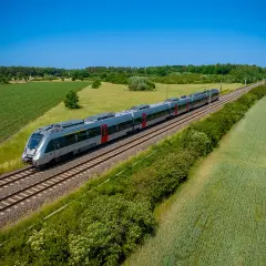 Moderner Triebzug fährt auf einer **railway deutsch** durch grüne Felder und Wälder unter blauem Himmel.