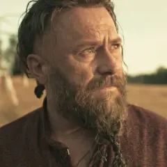 Brodaty mężczyzna z warkoczem na brodzie, jak z filmów Macieja Kawulskiego, spogląda w dal na tle pola.