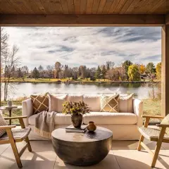 Cozy lakehouse interior design with a white sofa, chairs, and coffee table on a covered patio overlooking a serene lake and autumn foliage.
