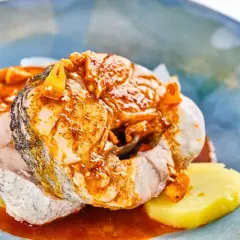 Rodajas de pescado blanco cubiertas con una sabrosa salsa gallega, servidas sobre patatas cocidas en un plato azul.