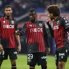 Piłkarze OGC Nice w czarno-czerwonych strojach dyskutują na boisku. Jeden z nich, z numerem 29, wskazuje palcem.