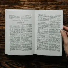 Dłonie trzymają otwartą księgę, jedną z apostolskich ksiąg Nowego Testamentu, obok leży notatnik i kubek kawy.