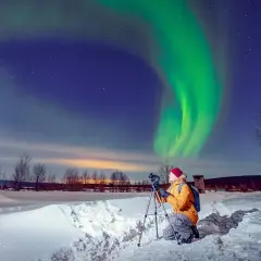 Fotograf uwiecznia zorzę polarną podczas zimowej wyprawy. Zielona poświata tańczy na nocnym niebie nad ośnieżonym krajobrazem.