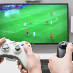 Dwie osoby grają w gry sportowe na konsoli, trzymając kontrolery przed ekranem telewizora z boiskiem piłkarskim.