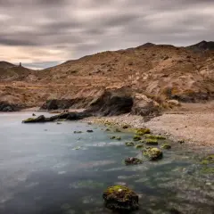 Mar tranquilo en la cala invencible Almería, con rocas cubiertas de algas y ruinas en la ladera.