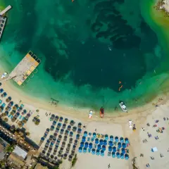 Widok z lotu ptaka na plażę z rzędami parasoli i leżaków, turkusową wodą i łodziami. Idealne miejsce na wakacje w albańskich miastach nad morzem.