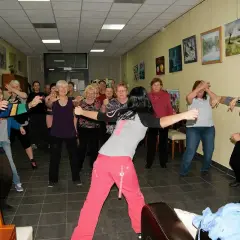 Grupa seniorów ćwiczy w sali Domu Seniora Zielony Zakątek. Wszyscy uśmiechnięci, w ruchu, w tle obrazy.
