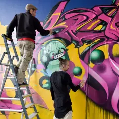 Drei Künstler sprühen bunte Graffiti auf eine Wand, einer auf einer Leiter.