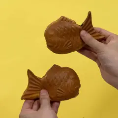 Zwei goldbraune Taiyaki, beliebte japanische Desserts in Fischform, werden von Händen auf gelbem Hintergrund gehalten.