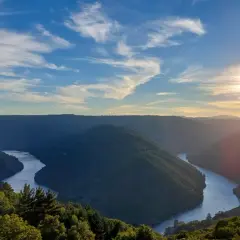 Un río serpentea entre montañas verdes, bajo un cielo azul con nubes finas. El sol se pone, tiñendo el horizonte de dorado. Parece el cabo do mundo.