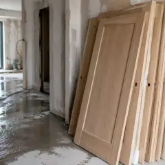 Portes en bois empilées, prêtes à être installées. Le sol est mouillé, signe que les travaux sont en cours pour empêcher le bois de travailler.