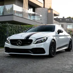 Biały Mercedes-AMG C63 Coupe z czarnymi pasami na masce, zaparkowany na brukowanej drodze przed nowoczesnym budynkiem.