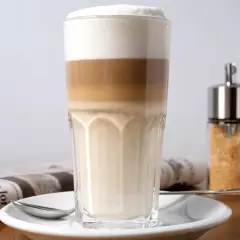 Ein geschichteter Latte Macchiato im Glas, daneben eine Zeitung und ein Zuckerspender. Milchkaffee vs. Latte Macchiato – die Wahl ist schwer!