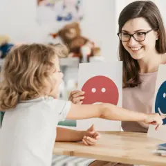 Dziecko wskazuje na uśmiechniętą buźkę, rozwijając inteligencję intrapersonalną. Terapeuta z uśmiechem prezentuje karty emocji.