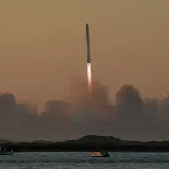Starship start. Rakieta wznosi się w niebo, zostawiając za sobą kłęby dymu. Na wodzie widać łodzie obserwujące ten spektakularny widok.