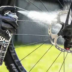 Gloved hand sprays Muc-Off Disc Brake Cleaner onto a bike's rotor, demonstrating how to clean up brake fluid for optimal performance.