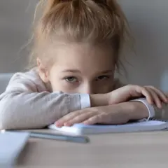 Ein trauriges Mädchen mit hochgesteckten Haaren liegt mit den Armen auf einem Notizbuch. Sie blickt mit müden Augen nach vorne, die schlechte Noten widerspiegeln.