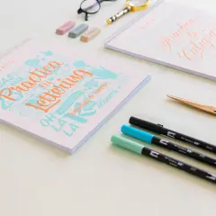 Cuadernos de "Practica Lettering" y "Practica tu Caligrafía" con rotuladores y tijeras doradas.