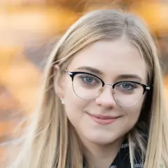 Uśmiechnięta blondynka w okularach, z rozmazanym, żółtym tłem. Idealna ogniskowa do portretu, podkreślająca oczy.